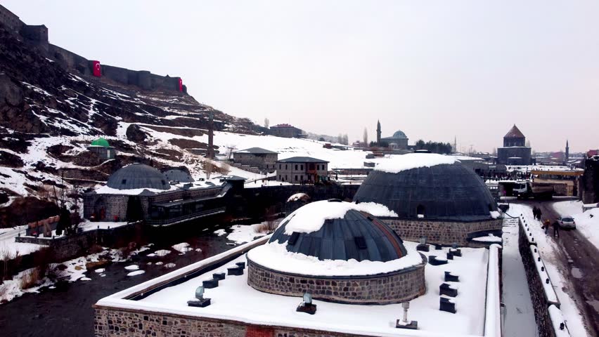 Dramatic drone footage of the historic Kars Castle and surrounding ancient stone structures blanketed in white snow during a cold winter day in Eastern Anatolia