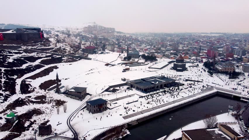 Panoramic aerial view of a modern city square and riverfront park covered in deep snow, showcasing the winter atmosphere of Kars city from above