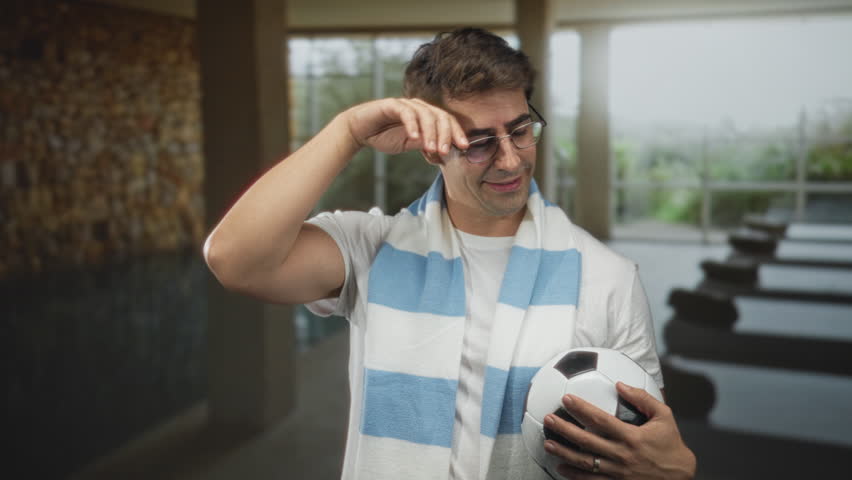 Man holding soccer ball, smiling and wearing a light blue and white scarf in a building lobby; pride camaraderie.