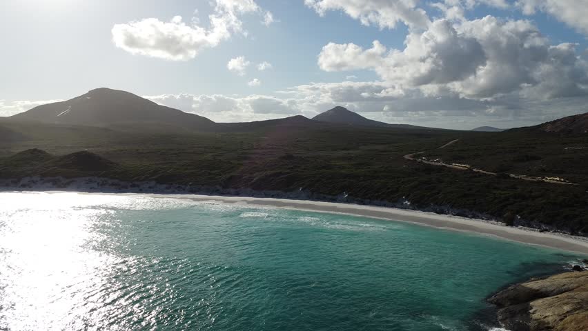Cinematic drone footage flying from the turquoise waters of Hellfire Bay toward the shoreline in Cape Le Grand National Park, Western Australia, with Frenchman Peak visible in the distance. Scenic coastal landscape combining crystal clear ocean, white sand and iconic granite mountain backdrop.