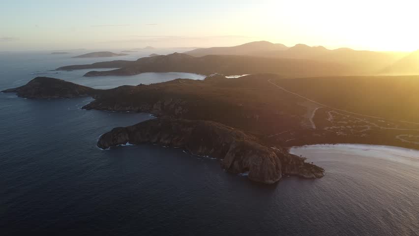 Ultra-wide aerial drone footage capturing Cape Le Grand National Park near Lucky Bay at sunset, Western Australia. Expansive panoramic landscape showcasing rugged coastline, turquoise bays and golden light over the remote Australian wilderness. Epic scenic establishing shot of pristine national park coastline.