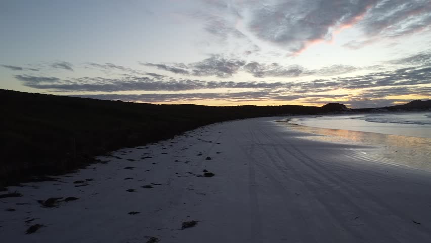 Cinematic drone footage captured at sunrise over Lucky Bay in Cape Le Grand National Park, Western Australia. Soft morning light illuminating turquoise waters and pristine white sand from a beach-level aerial perspective, creating a peaceful and atmospheric Australian coastal landscape.