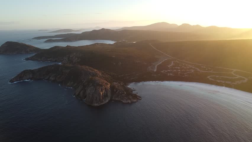 Ultra-wide aerial drone footage capturing Lucky Bay at sunset in Cape Le Grand National Park, Western Australia. Expansive panoramic view of brilliant white sand, turquoise waters and rugged coastline illuminated by warm golden hour light. Iconic Australian beach landscape from above.