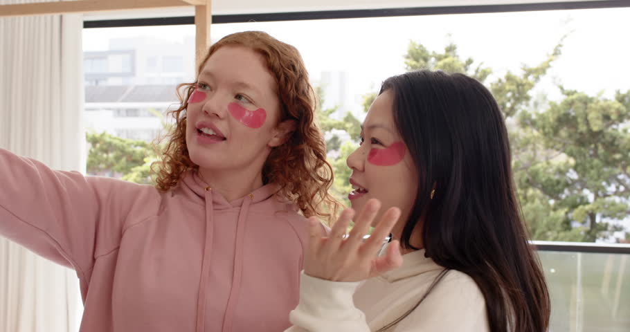 Diverse female friends at window, left woman pointing, other showing pink patches for skincare. Duo, hydrogel, daylight, balcony, candid, beauty, lifestyle
