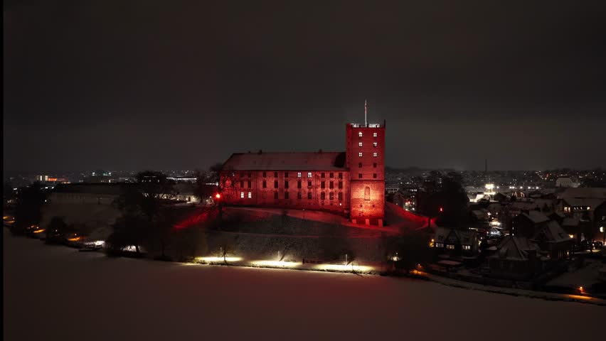 Cinematic aerial drone orbit around Koldinghus castle in Kolding, Denmark during a calm winter night. The historic medieval Danish landmark is beautifully illuminated in red light, standing prominently against the dark Nordic sky. A thin, even layer of seasonal snow covers the surrounding lake, creating a soft, textured foreground that enhances the atmospheric mood of the scene.

Filmed with a smooth GPS-controlled flight path, the camera movement is steady and consistent, making this footage ideal as a professional establishing shot. The dramatic lighting and tranquil winter environment provide strong visual contrast between the warm architectural illumination and the cold Scandinavian night landscape.

This aerial footage is suitable for documentary productions, historical programs, travel features, tourism promotion, cultural storytelling, climate or seasonal content, and cinematic background visuals. The scene conveys themes of heritage, history, Nordic identity, winter atmosphere, and European architecture.

No people are visible. No logos or trademarks present. Clean composition with natural ambient city lights in the distance. High resolution drone footage with stable motion and professional framing.

Ideal for use in television, streaming productions, corporate films, promotional media, or editorial storytelling focused on Denmark, Scandinavian landmarks, medieval history, winter environments, or atmospheric night visuals.
