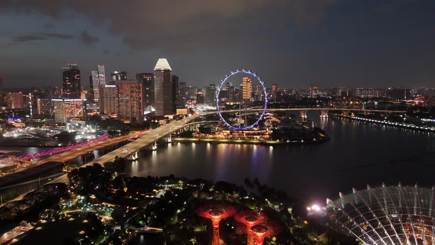 Singapore by Night – 4K aerial drone footage of marina bay sands, city skyline, skyscrapers, luxury hotels, gardens by the bay, garden Rhapsody, dome and river illuminated, Flyer (sun wheel) in financial district.