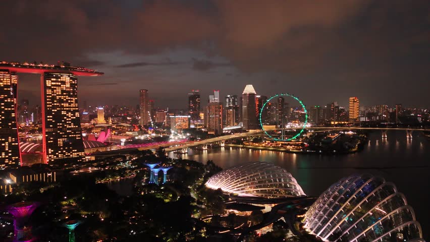 Singapore by Night – 4K aerial drone footage of marina bay sands, city skyline, skyscrapers, luxury hotels, gardens by the bay, garden Rhapsody, dome and river illuminated, Flyer (sun wheel) in financial district.