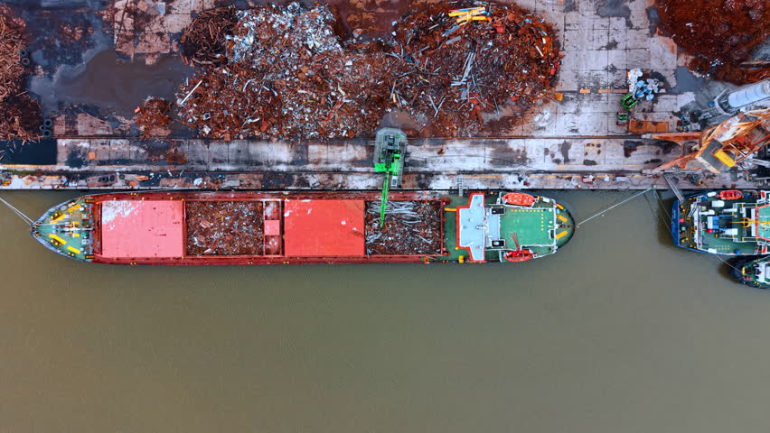Aerial view of cargo ship loading process at shipyard. A red vessel at dock with green crane and large piles of industrial scrap metal nearby.