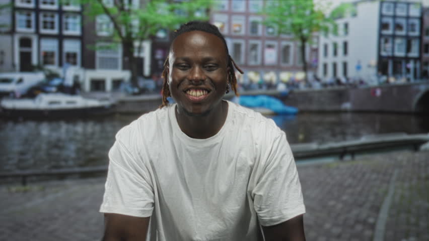 Man showing thumbs up and smiling on a street by an amsterdam canal with boats and water visible; joy celebration.