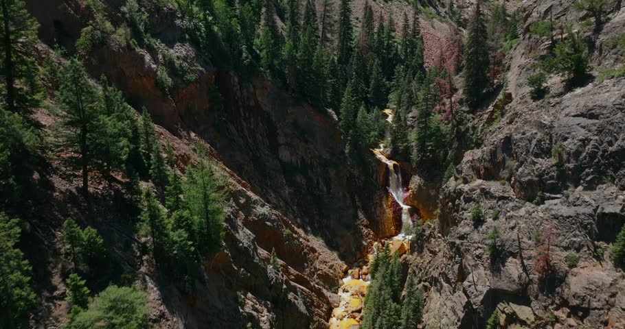 High angle aerial of hidden roadside waterfall flowing by rugged alpine canyon terrain, Million Dollay Highway Colorado USA