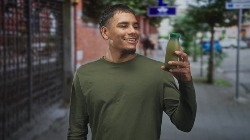 Man smiles while holding green juice bottle with hand raised on a city street near a brick wall; friendly wellness.
