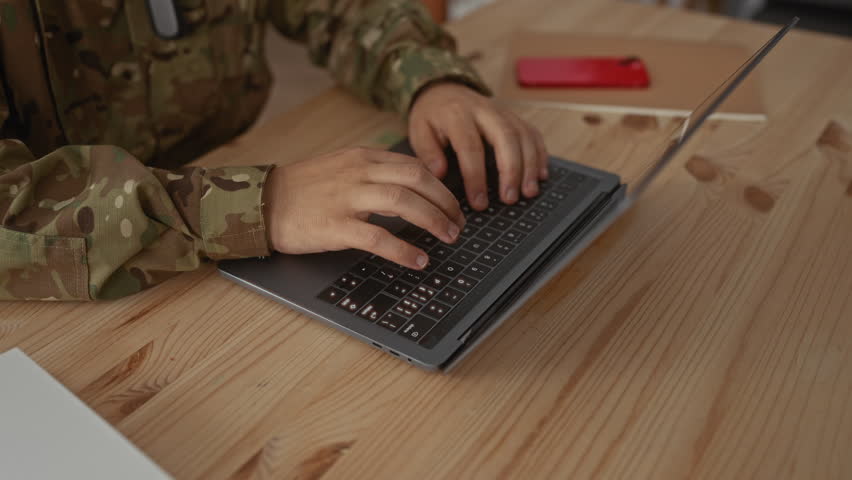Young hispanic man in camouflage uniform typing on laptop on wooden table in building interior; determination.