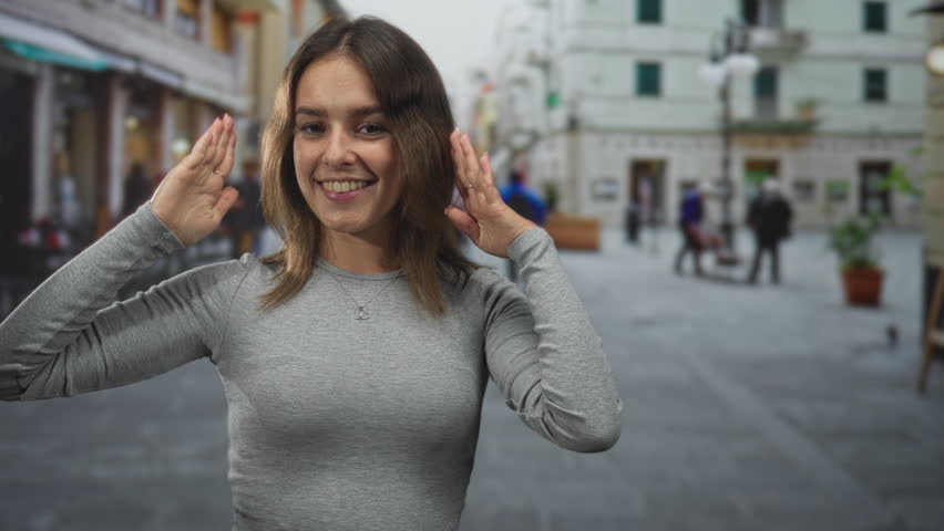 Woman smiling and posing with hands framing her face and palms up on a cobblestone street plaza in the city; joy travel.