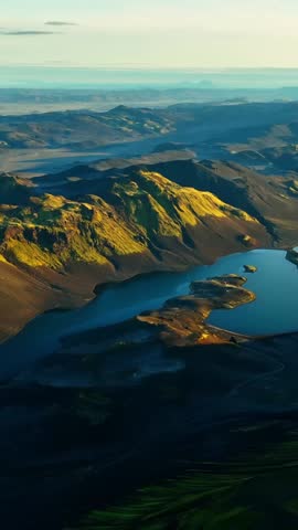 Vertical drone view of Iceland highlands: blue volcanic lake beside moss-covered mountains and black lava sands at golden hour, with wide sky copy space.