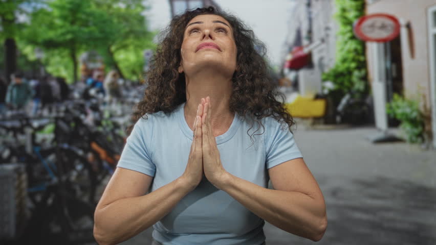 Young hispanic woman pressing hands in prayer on street surrounded by parked bicycles under sunlight; serenity.