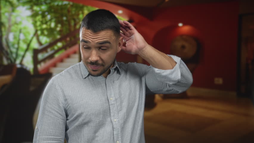 Young hispanic man casually touching neck while standing in a hotel building lobby with wooden staircase in view; uncertainty.