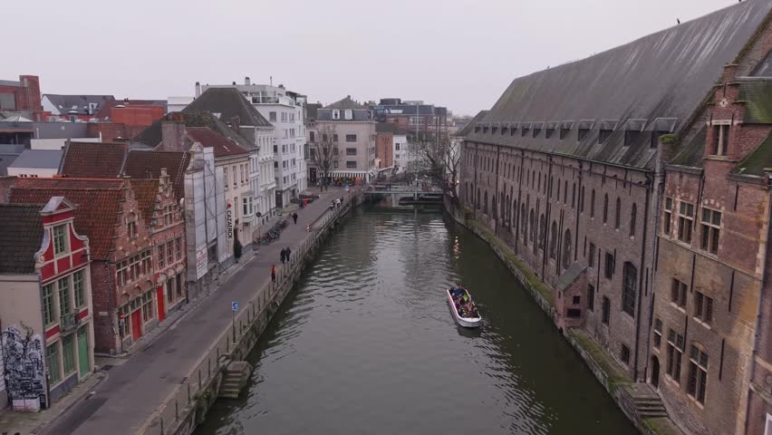 Drone pulls back from a tourist boat near Saint Michael’s Church, revealing the iconic bridge and the stunning medieval cityscape of Ghent along the River Leie.