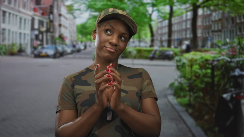 Woman in camouflage uniform clasps dogtags and folds hands on a street with parked cars and brick buildings; readiness determination.
