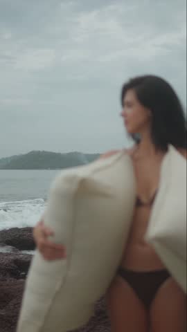 Woman in swimwear holding large soft cushions while standing on rocky beach near ocean waves under cloudy sky, calm coastal lifestyle scene.
