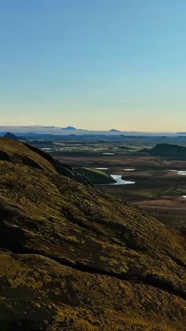 Drone view from a mossy mountainside over Iceland highlands plain with a winding river and distant crater lakes under vast clear sky, warm evening light for cinematic travel b-roll.