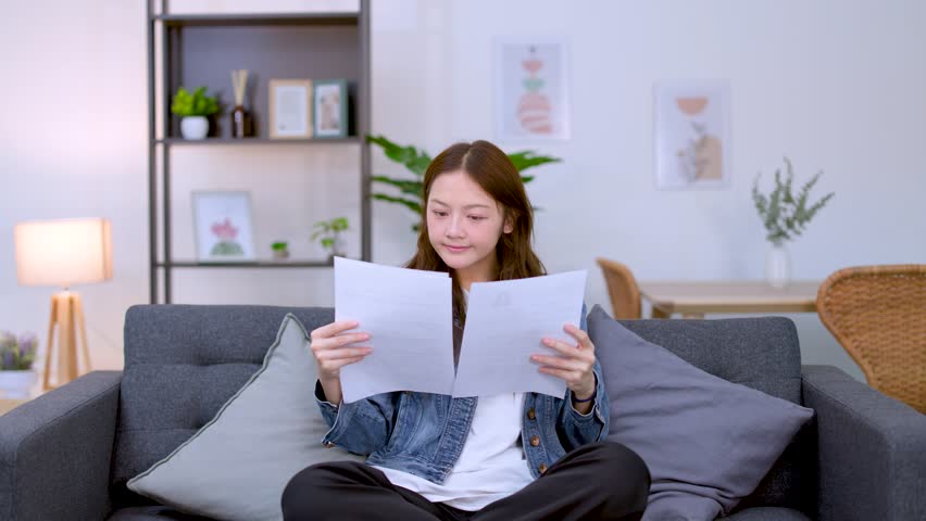 A young woman sits on a sofa, carefully reading and comparing documents with focused expressions