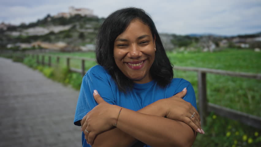African american woman gently hugging her arms on a street walkway by a green field and wooden fence; serenity.