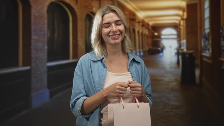 Woman holding a pink shopping bag with both hands clasped at waist, smiling and glancing upward in an indoor building corridor lined with arches and warm lights; serenity leisure.
