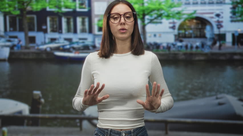 Young woman wearing glasses holds both hands in an ok gesture on a street beside a canal and moored boats; calm contemplation.