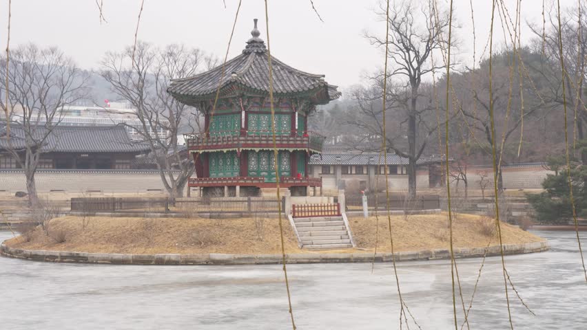 Slow motion landscape of Hyangwonjeong Pavilion hut temple building on island surrounded by frozen lake and trees with reeds in foreground at Gyeongbokgung Palace grounds Seoul South Korea Asia travel