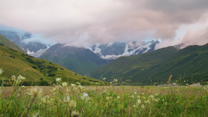 Georgia Svaneti Caucasus mountains spring zoom in timelapse, low clouds roll across alpine valley, snow peaks and glaciers, green hills, wildflower meadow foreground, dramatic weather shift