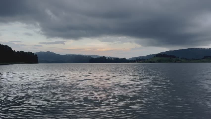 Boat-level view across Lake Bunyonyi near Kabale, southwestern Uganda, showing rippled lake surface, distant terraced hills and storm clouds, peaceful travel mood