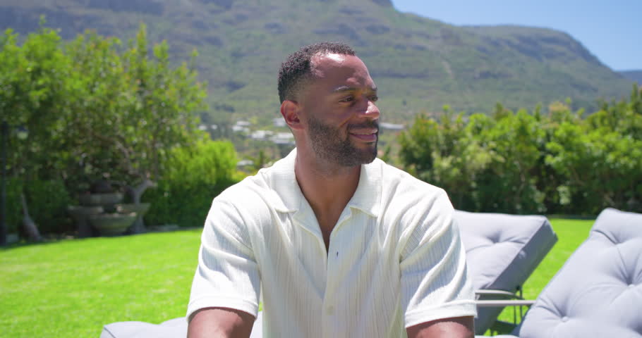 Mature man sitting on cushioned chair on backyard lawn reacting to off camera voice, smiling. Outdoor, garden, patio, lounge, mountains, hedge, greenery
