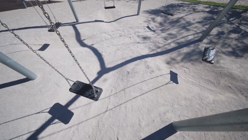 A close-up perspective of empty swings and their geometric shadows stretching across the sandy ground of a public park.