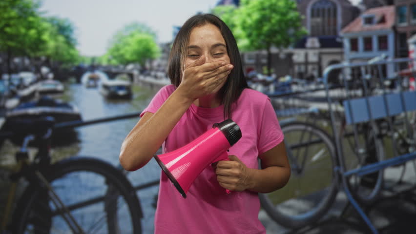 Woman covering mouth with hand while holding a pink megaphone on street in amsterdam; amusement spontaneity energy.