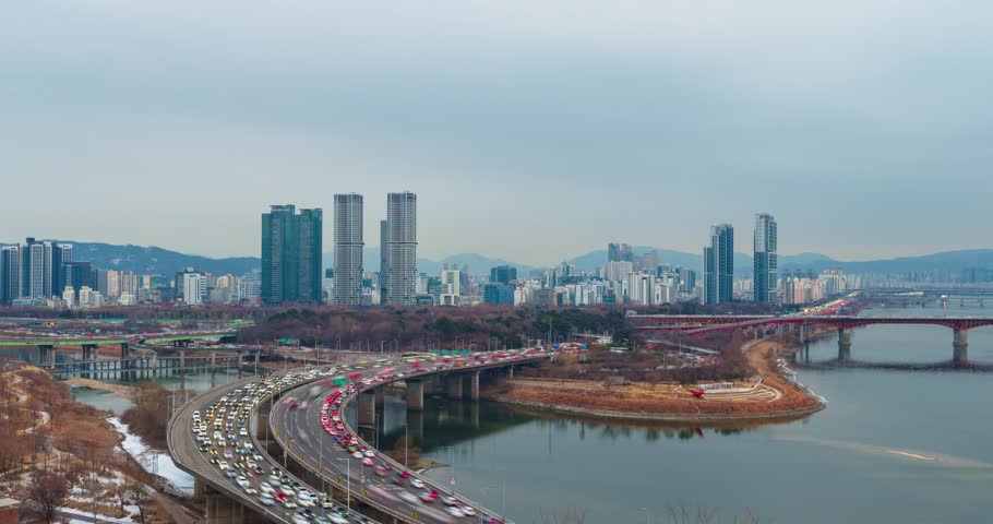 Seoul day to night hyperlapse zoom out, heavy traffic on Gangbyeonbuk-ro expressway, Seongsu Bridge, Han River, Lotte Tower, twin towers skyline, mountains, winter overcast transitioning to blue hour