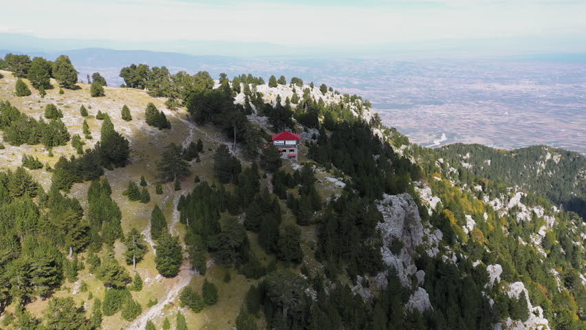 Slow panning drone shot of Spilios Agapitos Refuge nestled among fir trees in the rocky landscape of Mount Olympus under a blue sky with clouds