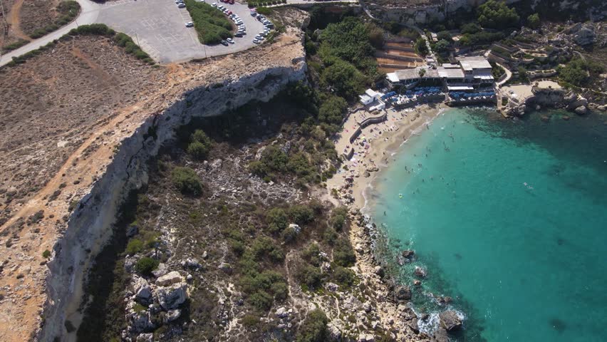 Wide aerial shot of Paradise Bay beach with parking lot, sandy beach, beach club, dramatic limestone cliff formations, and turquoise Mediterranean waters.