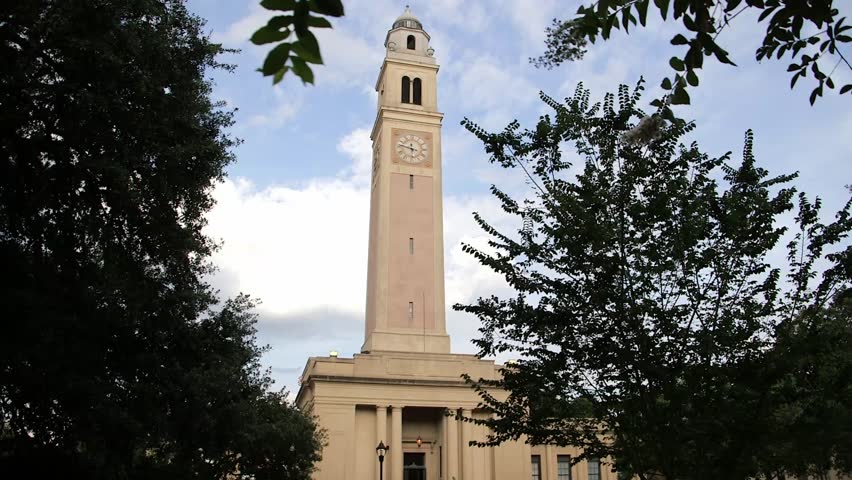 Iconic clock tower of Memorial Tower at Louisiana State University (LSU) campus in Baton Rouge, Louisiana, USA, neoclassical architecture with trees in foreground under blue sky.
