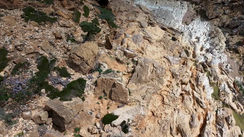 Aerial view of rugged rocky hillside with an eroded limestone ravine or dry creek bed, scattered green shrubs, and natural rock pool at the end of it. Sokotra island.