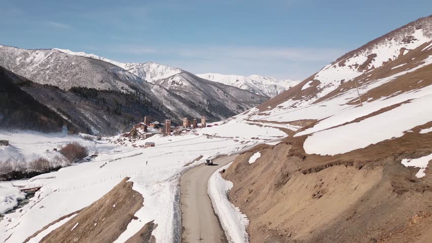 Aerial view Ushguli Svaneti mountain road, winter snowbanks, Caucasus peaks, remote village access, scenic road trip, adventure travel, alpine landscape, rural infrastructure