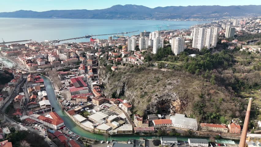 Drone aerial flying above the Rječina River through Rijeka toward the Kvarner Bay and the Adriatic Sea on a sunny day. Cityscape spreading below with coastline and bay in the distance.
