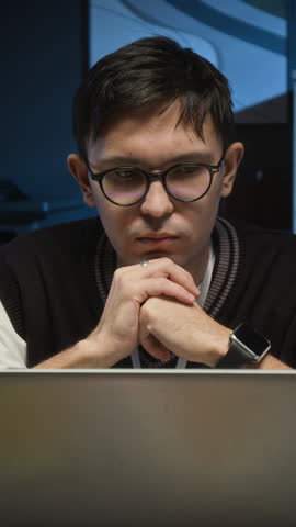 Vertical shot of focused young male project manager with glasses and wristwatch working on laptop in dimly lit office
