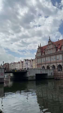 Historic brick granary building with red tiled roof along the waterfront in Gdansk old town, Poland, reflected in calm water under a dramatic cloudy sky.