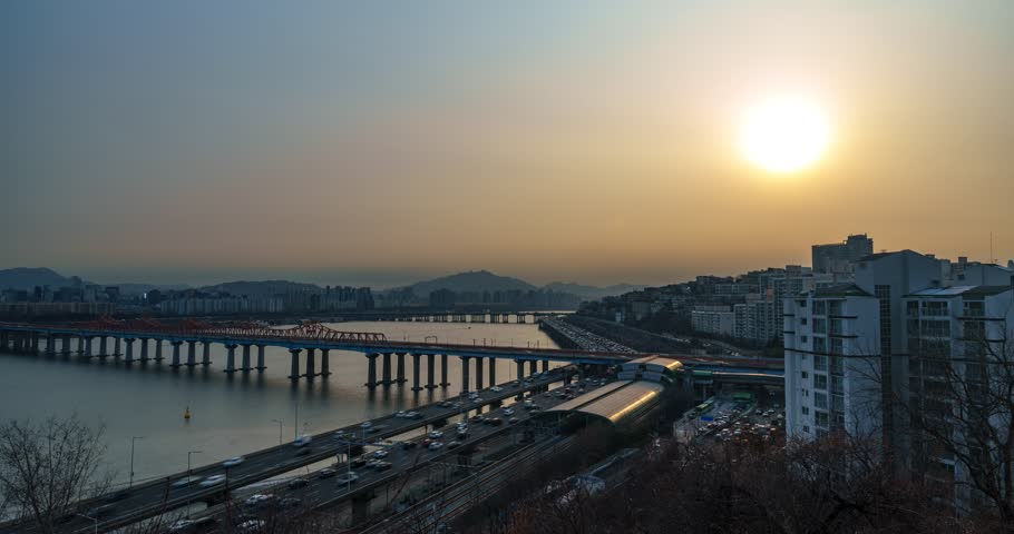 Seoul, South Korea time lapse, traffic on Gangbyeonbuk-ro expressway, Donho Bridge, Oksu Station, Han River reflecting golden sunset, silhouetted mountains, winter haze, dramatic orange sky