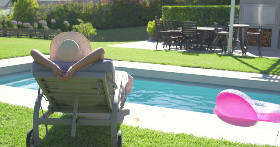 African American woman in straw-hat relaxing in sun on lounge, reaching pool as pink float drifting. Backyard, outdoor, private, relaxation, sunlit, inflatable, garden