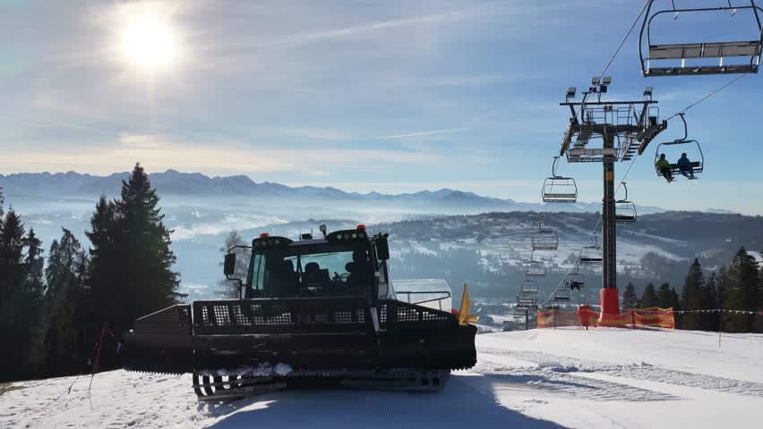 Snow grooming machine preparing ski slope near Zakopane with chairlift and Tatra Mountains in background, winter resort scene showing piste maintenance, alpine landscape and clear sunny day	
