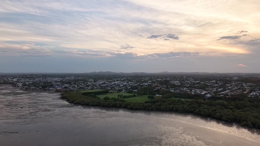 Aerial view of a coastal industrial area featuring large storage tanks and factory buildings situated between a dense mangrove forest and a muddy riverbed under a cloudy evening sky