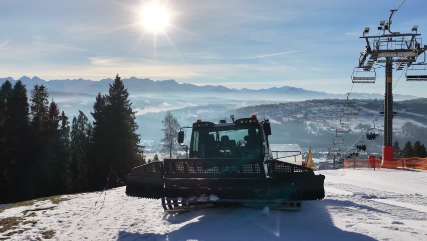 Snow grooming machine preparing ski slope near Zakopane with chairlift and Tatra Mountains in background, winter resort scene showing piste maintenance, alpine landscape and clear sunny day	
