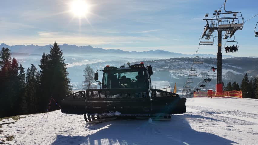 Snow grooming machine preparing ski slope near Zakopane with chairlift and Tatra Mountains in background, winter resort scene showing piste maintenance, alpine landscape and clear sunny day	
