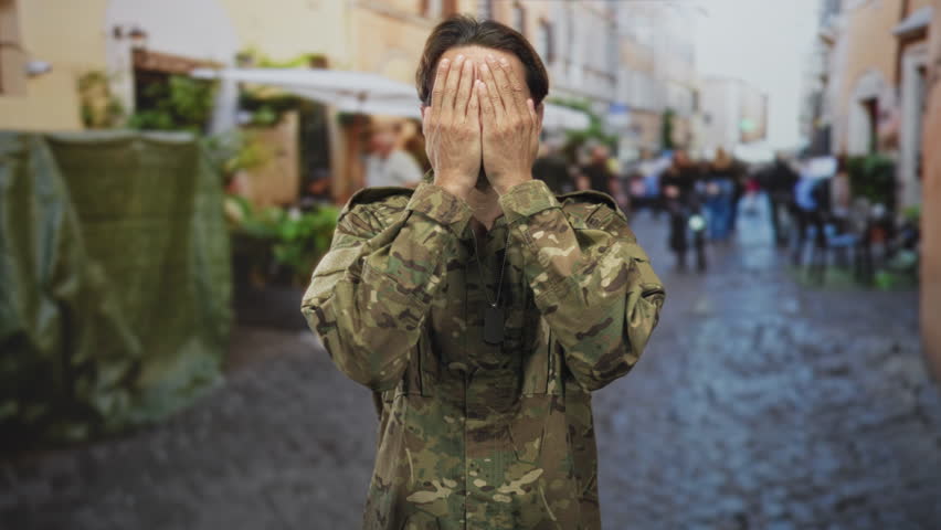Soldier man in camouflage covers face with hands on a cobbled street near blurred cafe diners and passing cyclists; playful resilience duty.